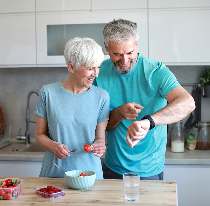 Smiling older couple in kitchen preparing fruit and checking smartwatch during ZepBound monthly progress check-in.