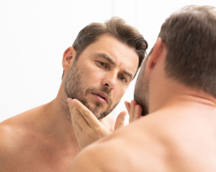 Shirtless man examining face in mirror, highlighting common men’s skincare issues like irritation and acne.