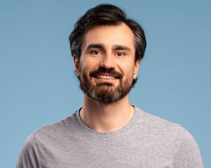 Smiling bearded man in gray shirt against blue background, representing healthy skin and proper beard care.