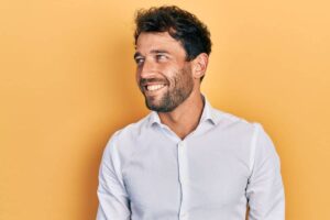 Smiling man in white shirt standing against yellow background, representing insurance coverage for rhinoplasty.