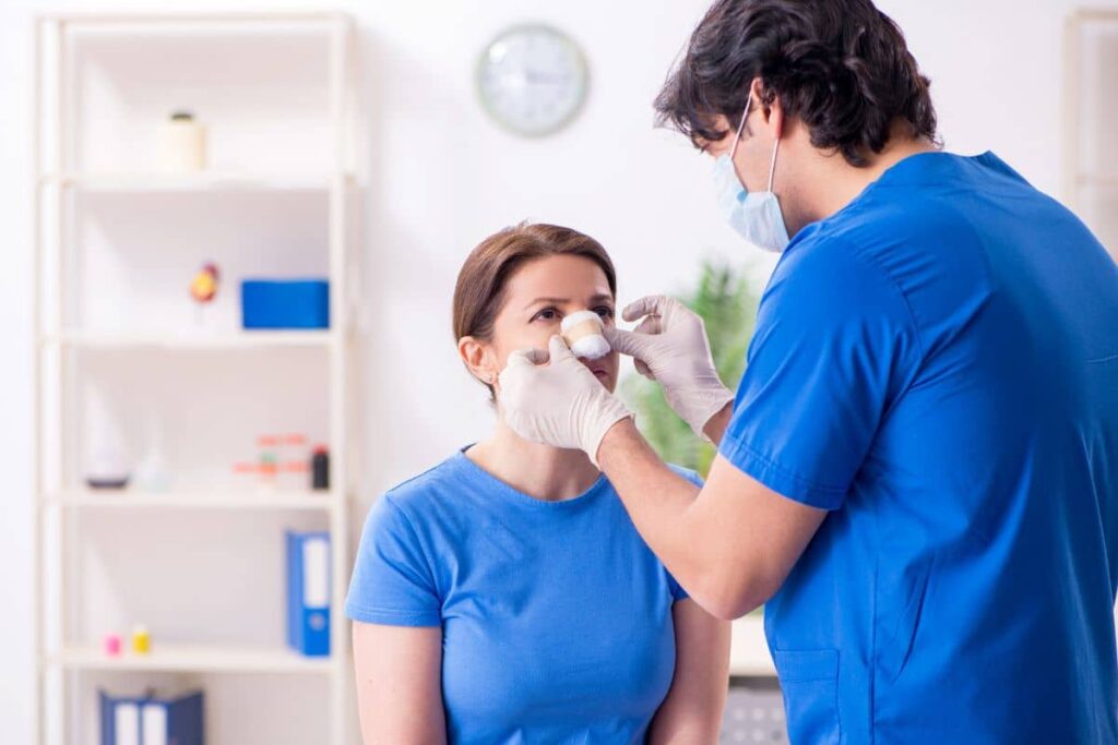 Doctor in blue scrubs providing post-operative nasal care to female rhinoplasty patient in medical office setting.