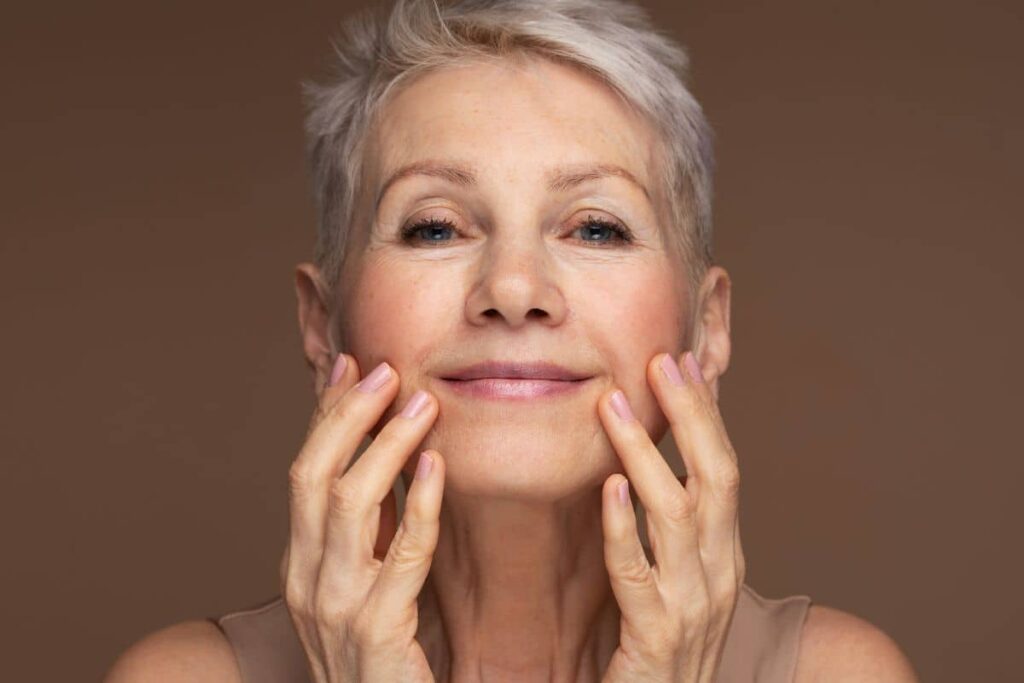 Older woman with short gray hair smiling gently, touching her face to highlight age-related facial symmetry changes.