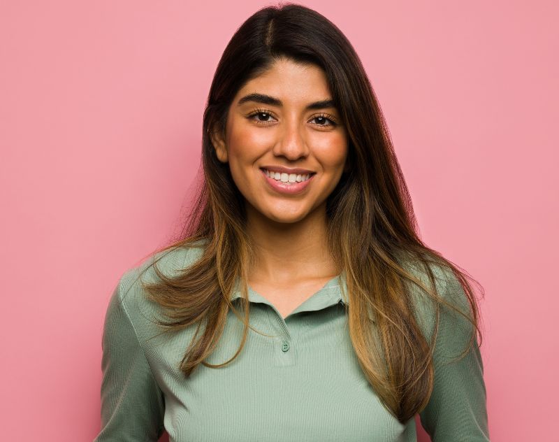 Confident Latina woman smiling against pink background, symbolizing Spanish-speaking support in rhinoplasty consultations.