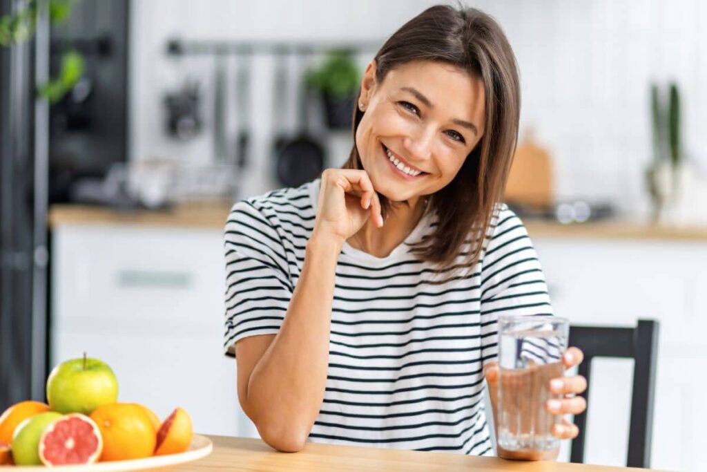 Smiling woman drinking water at kitchen table, preparing for a Botox or filler appointment with healthy habits.