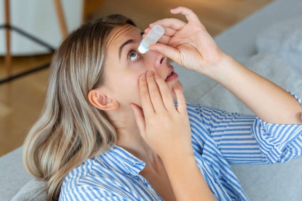 Woman applying eye drops to relieve dry eyes after blepharoplasty surgery while resting at home.