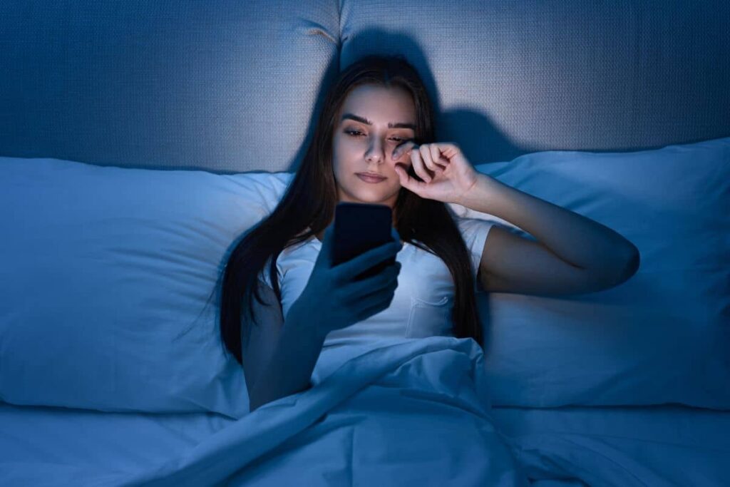 Woman lying in bed using smartphone, rubbing eyes from screen strain during eyelid surgery recovery.