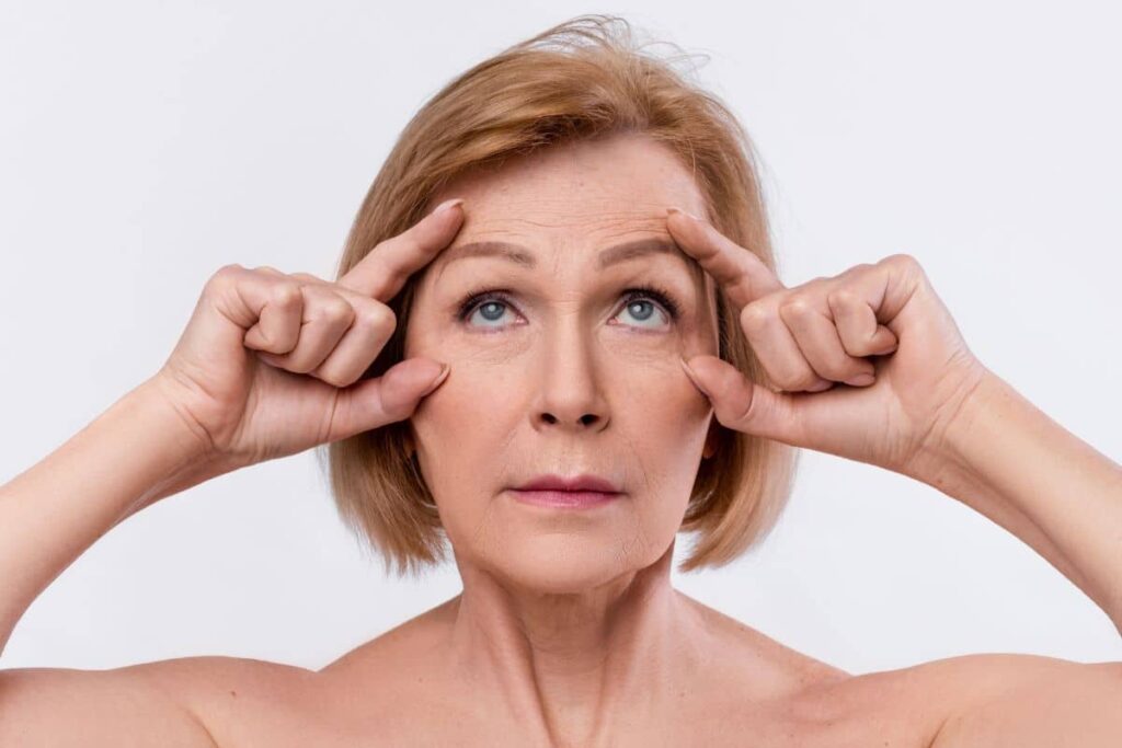 Mature woman demonstrating brow lift effect by raising forehead skin to reduce wrinkles and lift sagging brows.