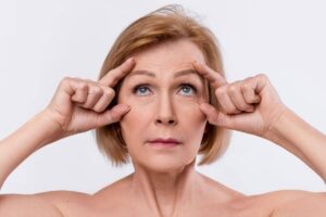 Mature woman demonstrating brow lift effect by raising forehead skin to reduce wrinkles and lift sagging brows.