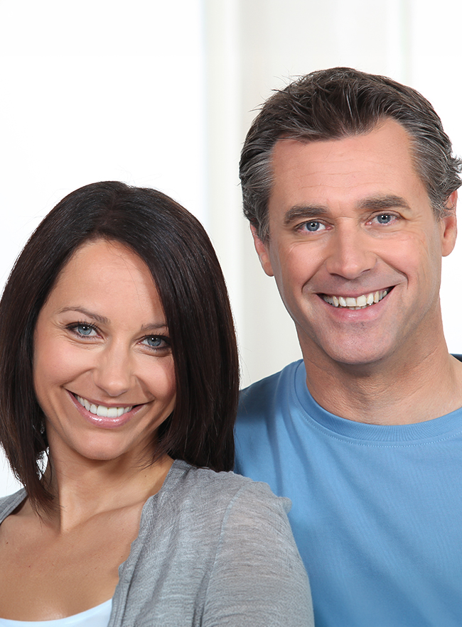 Smiling middle-aged couple with natural-looking facial rejuvenation, smooth skin, and relaxed expressions indoors.
