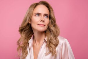 Woman with soft, natural facial features and minimal makeup against pink background, reflecting modern aesthetic refinement trend