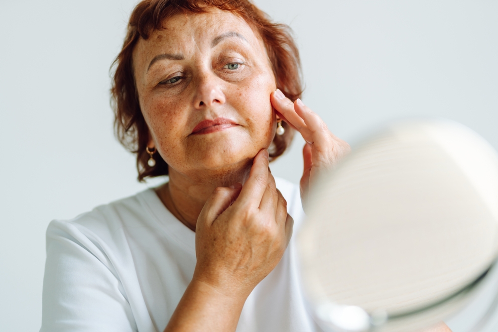 Older woman touching her face in mirror, illustrating facial bone resorption and age-related structural facial changes.