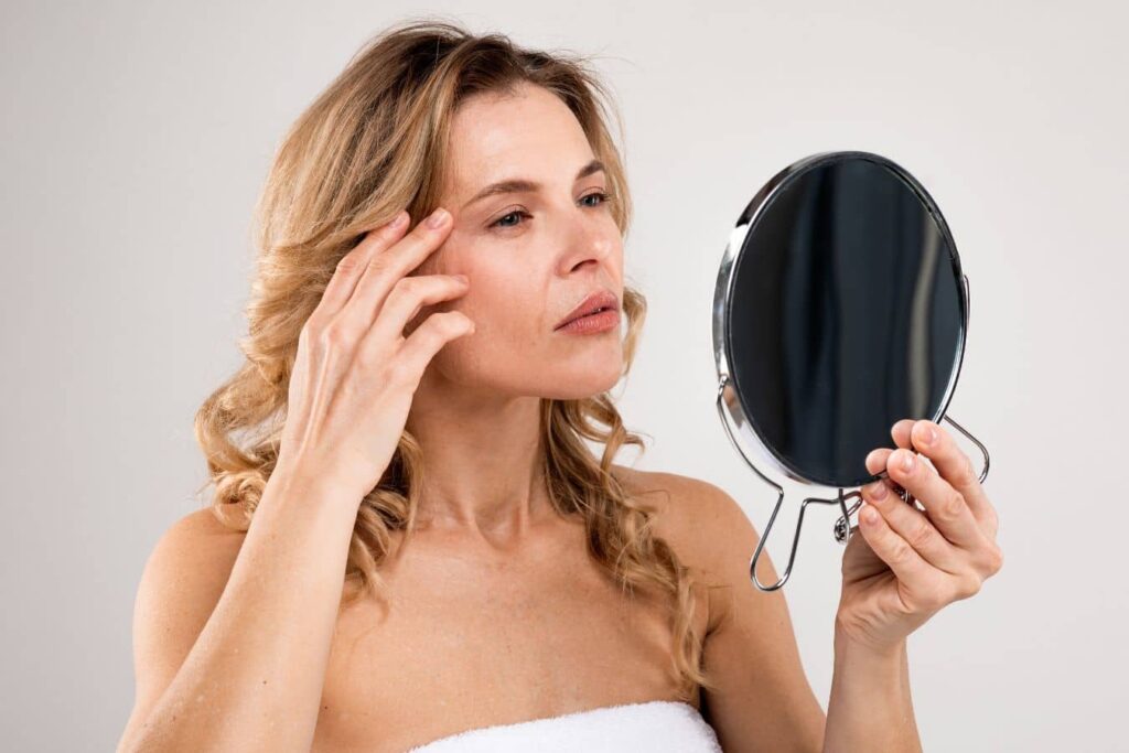 Woman examining her face in a mirror, illustrating facial aging, hollow cheeks, and shifting fat compartments.