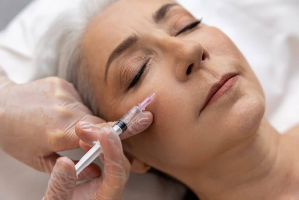 Practitioner injecting cheek filler into a mature woman, illustrating risks of facial overfilling and unnatural contours.