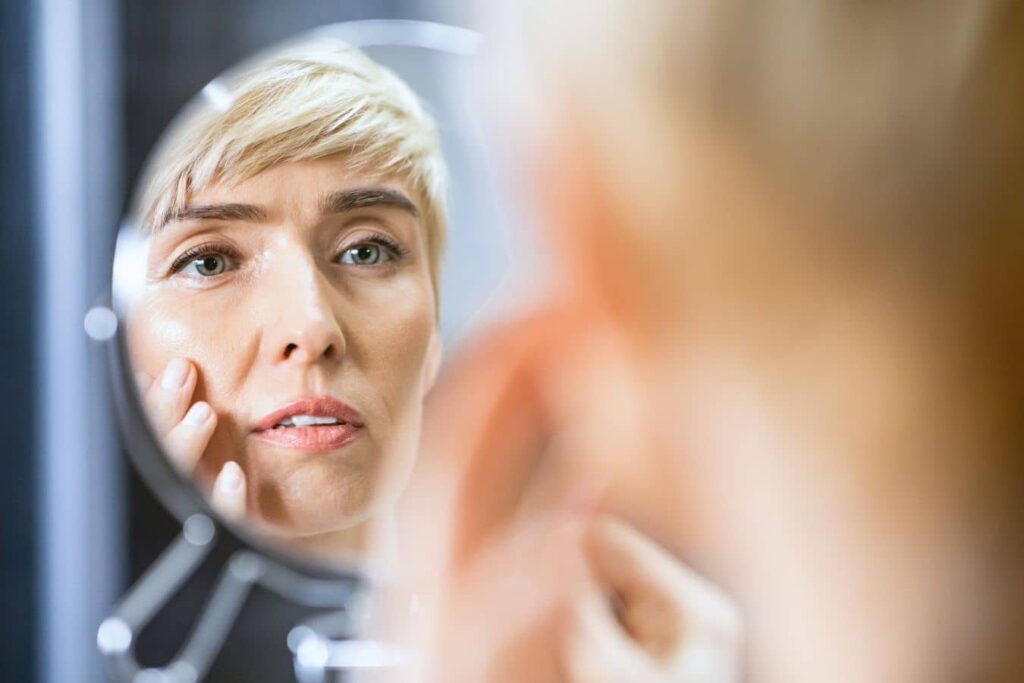 Woman examines early facial aging in a mirror, highlighting structural changes affecting the midface and jawline.